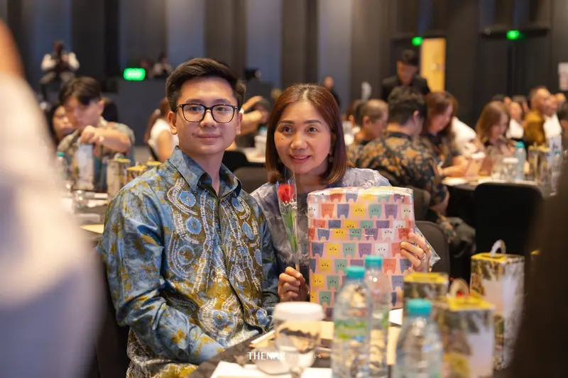 Pria muda berkacamata dan seorang ibu duduk berdampingan, sang ibu memegang bingkisan dan setangkai mawar
